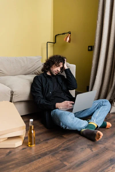 Enfoque selectivo del teletrabajo apuntando con el dedo a la computadora portátil cerca de cajas de cerveza y pizza en el suelo - foto de stock