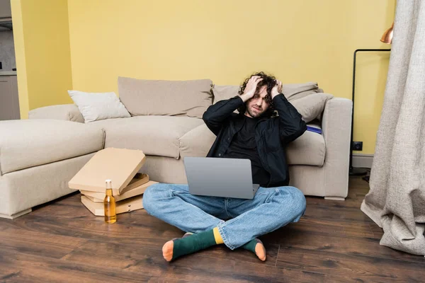 Exhausted man holding laptop while sitting on floor near beer bottle and pizza boxes — Stock Photo
