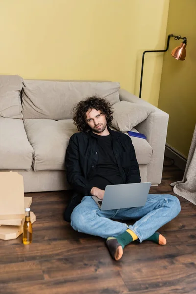 Enfoque selectivo del hombre guapo usando el ordenador portátil cerca de la botella de cerveza y cajas de pizza en el suelo en la sala de estar - foto de stock