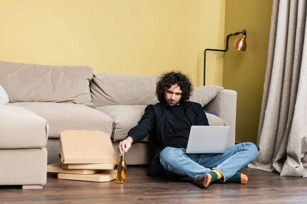 Freelancer holding bottle of beer near laptop and pizza boxes on floor in living room — Stock Photo