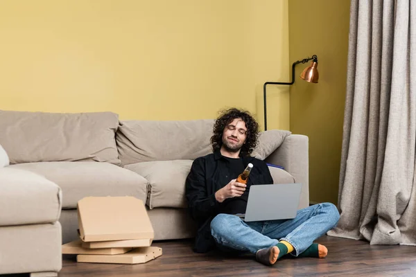 Focus selettivo dell'uomo che tiene la bottiglia di birra vicino al computer portatile e scatole di pizza sul pavimento a casa — Foto stock