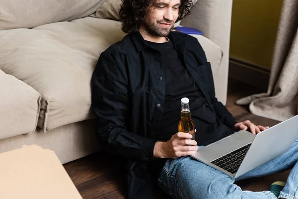 Selective focus of man holding beer bottle and using laptop near pizza box on floor — Stock Photo