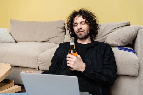 Focus selettivo di freelance in possesso di bottiglia di birra e utilizzando laptop vicino a scatole di pizza e divano a casa — Foto stock