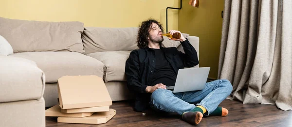 Panoramic shot of freelancer drinking beer near laptop and pizza boxes on floor — Stock Photo