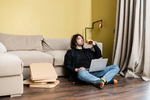 Selective focus of man drinking beer while using laptop near pizza boxes on floor — Stock Photo