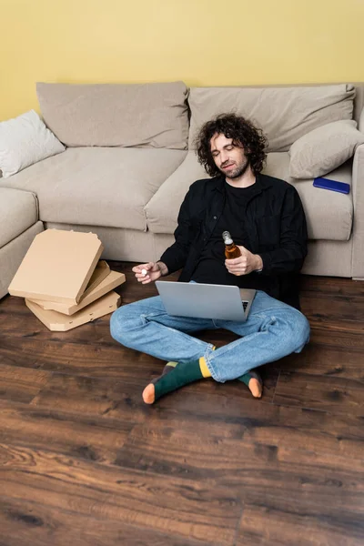 Curly teleworker segurando garrafa de cerveja e usando laptop perto de caixas de pizza na sala de estar — Fotografia de Stock