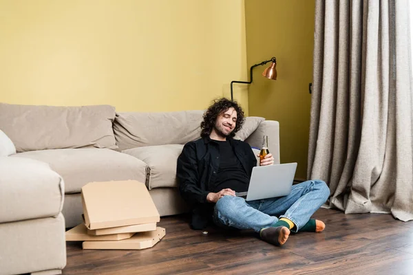 Selektiver Fokus eines lächelnden Mannes, der auf Laptop schaut und daheim eine Flasche Bier in der Nähe von Pizzakartons auf dem Boden hält — Stockfoto