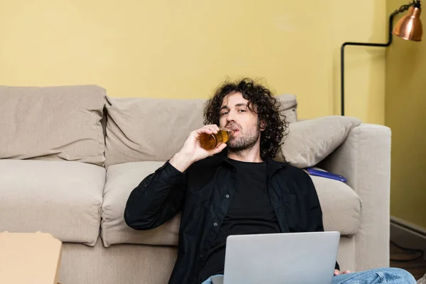 Красавчик смотрит в камеру и пьет пиво возле ноутбука и пиццу дома. — стоковое фото