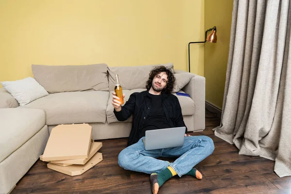 Curly man looking at camera while holding bottle of beer near laptop and pizza boxes on floor at home — Stock Photo