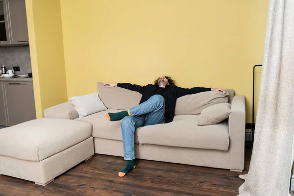 Hombre rizado mirando hacia arriba mientras está sentado en el sofá en la sala de estar - foto de stock