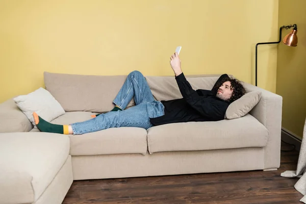 Vista lateral de homem bonito usando smartphone enquanto deitado no sofá em casa — Fotografia de Stock