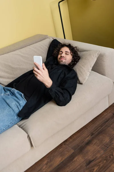 Lockiger Mann blickt in Kamera und hält Smartphone auf Couch — Stockfoto