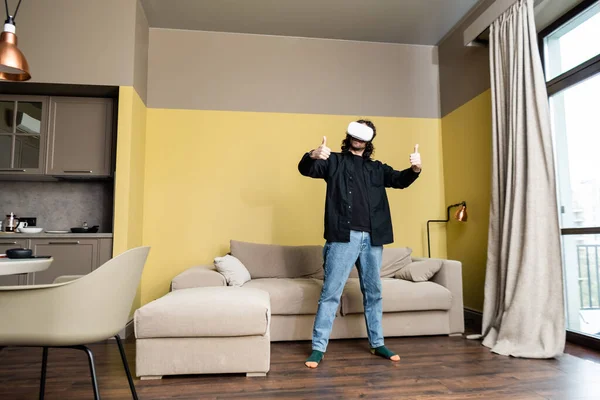 Hombre mostrando como gesto mientras usa auriculares de realidad virtual en la sala de estar - foto de stock