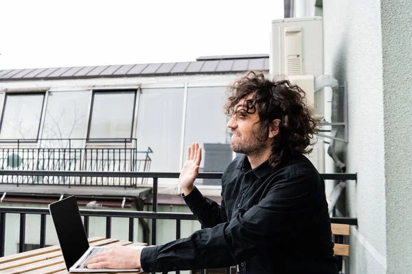 Side view of smiling teleworker waving hand at laptop on balcony — Stock Photo