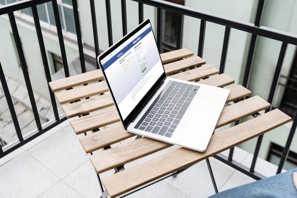 KYIV, UCRANIA - 25 de abril de 2020: Vista recortada del hombre sentado cerca de la computadora portátil con el sitio web de facebook en el balcón - foto de stock