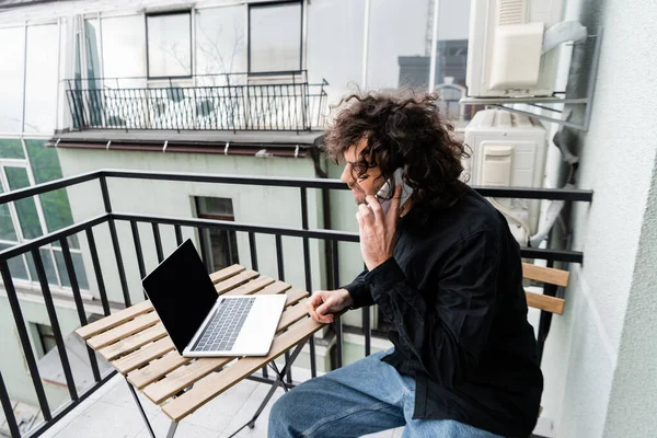 Teleworker talking on smartphone near laptop on table on balcony — Stock Photo
