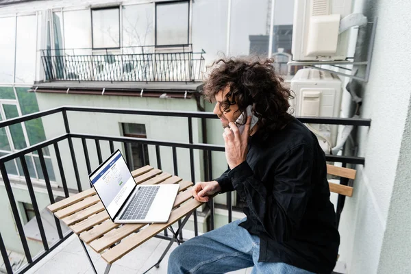 KYIV, UCRANIA - 25 DE ABRIL DE 2020: Teletrabajador hablando en el teléfono inteligente cerca de la computadora portátil con el sitio web de facebook en la mesa en el balcón - foto de stock