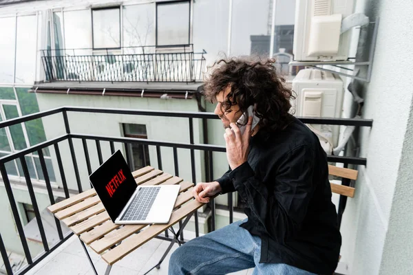 KYIV, UKRAINE - 25 AVRIL 2020 : Un homme bouclé parle sur un smartphone près d'un ordinateur portable avec le site web netflix sur le balcon — Photo de stock