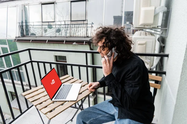 KIEW, UKRAINE - 25. APRIL 2020: Freelancer im Gespräch auf dem Smartphone in Laptopnähe mit Youtube-Website auf dem Tisch auf dem Balkon — Stockfoto