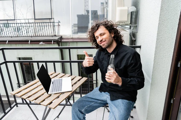 Homme souriant montrant comme geste tout en tenant bouteille de bière près d'un ordinateur portable sur le balcon — Photo de stock