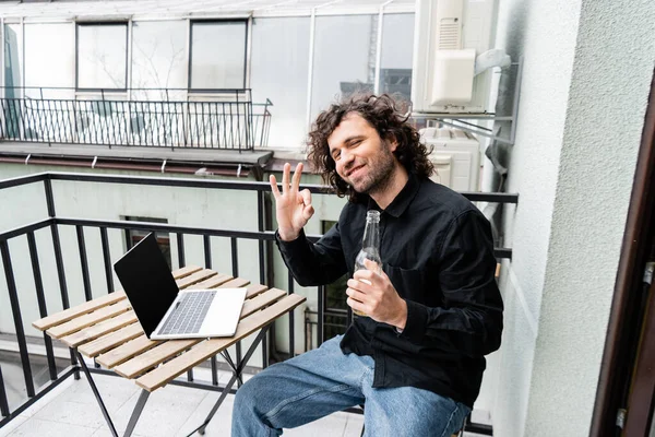Freelancer sorridente alla macchina fotografica e mostrando il gesto giusto mentre tiene la bottiglia di birra vicino al computer portatile sul balcone — Foto stock