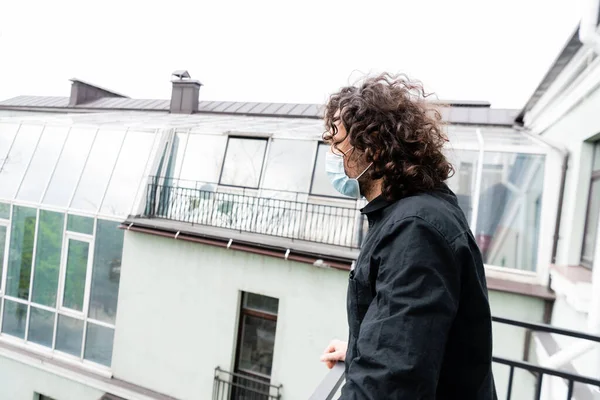 Side view of curly man in medical mask standing on balcony at home — Stock Photo