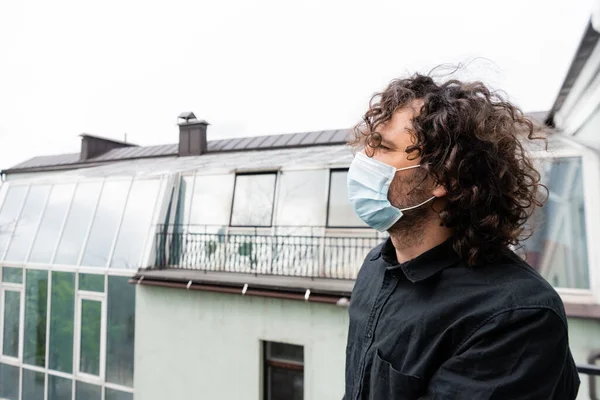 Вид сбоку на кудрявого человека в медицинской маске, смотрящего в сторону, стоя на балконе — стоковое фото