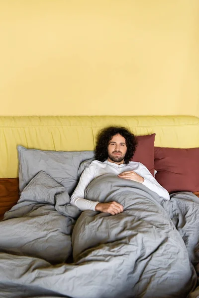 Lockiger Mann blickt in Kamera, während er auf dem Bett liegt — Stockfoto
