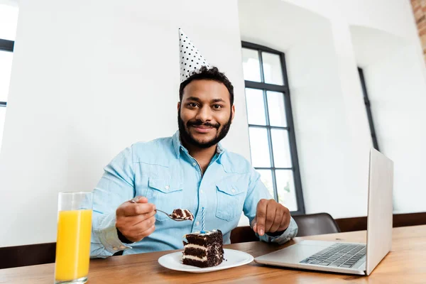 Felice freelance africano americano in cappello partito tenendo cucchiaio vicino gustosa torta di compleanno e laptop — Foto stock