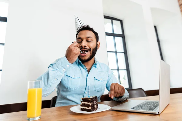 Africano americano uomo in partito cap mangiare gustoso compleanno torta vicino laptop — Foto stock