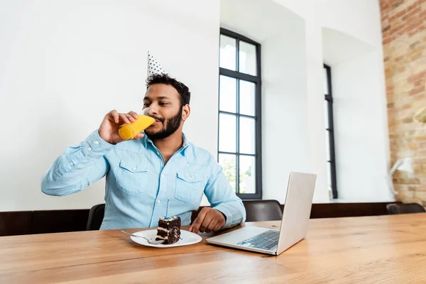 Afroamerikanisch-amerikanischer Freelancer in Partymütze trinkt Orangensaft in der Nähe von leckerem Geburtstagskuchen und Laptop — Stockfoto