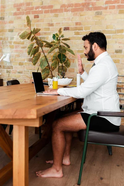 Barbudo hombre afroamericano en camisa y ropa interior sentado cerca de la computadora portátil y agitando la mano mientras tiene videollamada - foto de stock