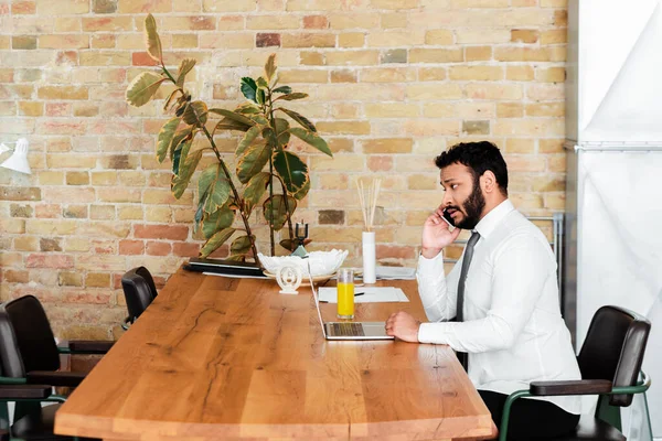 Freelancer americano africano barbudo em camisa sentado perto do laptop e falando no smartphone — Fotografia de Stock