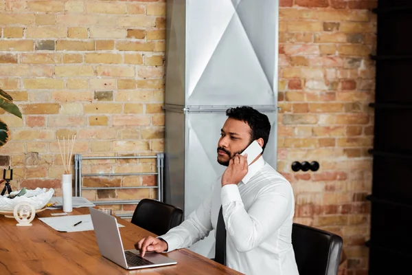 Bearded african american freelancer in suit sitting near laptop and talking on smartphone — Stock Photo