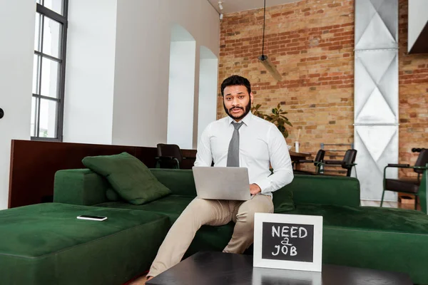 Homme afro-américain en utilisant un ordinateur portable près du tableau noir avec besoin d'un lettrage de travail et smartphone sur le canapé — Photo de stock