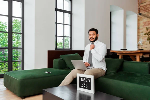 African american man in suit touching tie near laptop and chalkboard with need a job lettering — Stock Photo