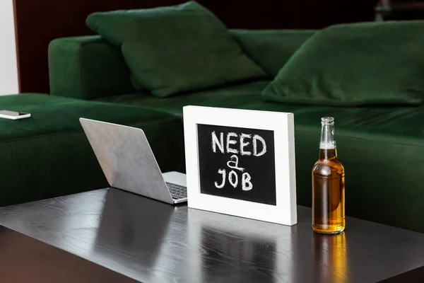 Laptop near chalkboard with need a job lettering and bottle of beer near sofa — Stock Photo