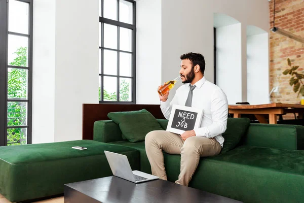 Unemployed african american man drinking beer and holding chalkboard with need a job lettering near gadgets — Stock Photo