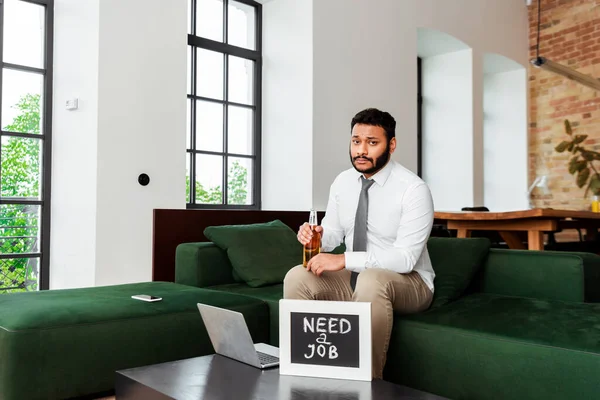 Unemployed african american man holding beer bottle near chalkboard with need a job lettering and laptop — Stock Photo