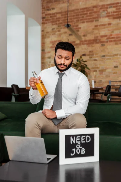 Desempregado afro-americano homem segurando garrafa de cerveja perto de quadro-negro com necessidade de um emprego lettering e laptop na mesa de café — Fotografia de Stock