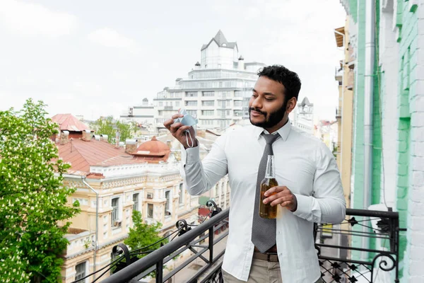 Bearded african american man holding medical mask and bottle of beer on balcony, end of quarantine concept — Stock Photo