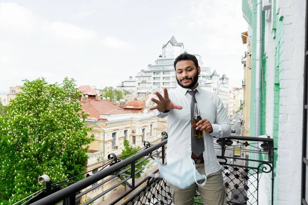 Messa a fuoco selettiva di uomo afroamericano barbuto gettando in maschera medica aria e tenendo bottiglia di birra sul balcone, fine del concetto di quarantena — Foto stock