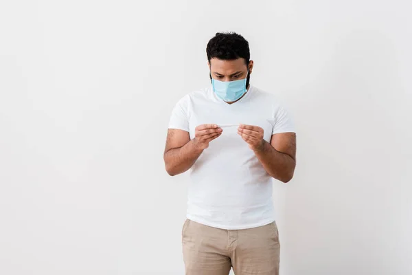 Hombre afroamericano en máscara médica mirando termómetro digital cerca de pared blanca - foto de stock