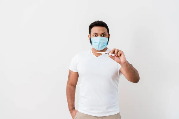 Homme afro-américain malade en masque médical debout avec la main dans la poche et tenant thermomètre numérique près du mur blanc — Photo de stock