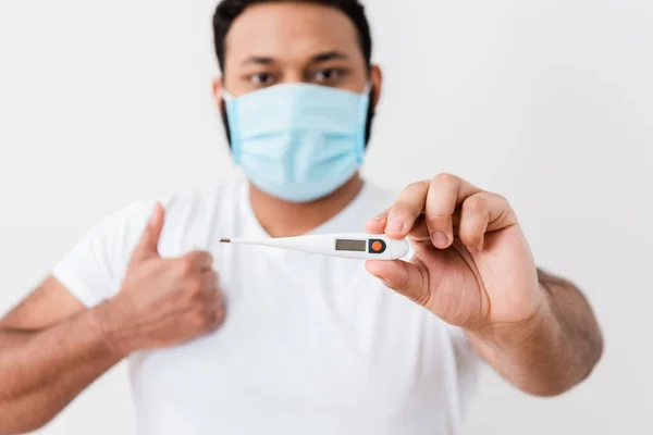 Enfoque selectivo del hombre afroamericano en máscara médica sosteniendo termómetro digital y mostrando el pulgar hacia arriba cerca de la pared blanca - foto de stock