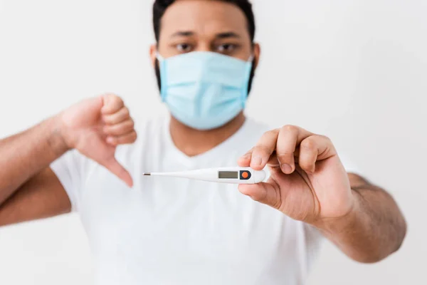 Enfoque selectivo del hombre afroamericano enfermo en máscara médica sosteniendo termómetro digital y mostrando el pulgar hacia abajo cerca de la pared blanca - foto de stock