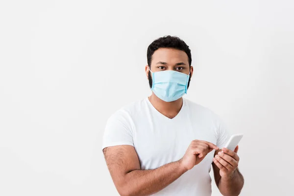 African american man in medical mask pointing with finger at smartphone near white wall — Stock Photo