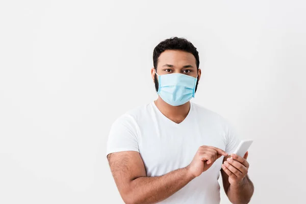 Hombre afroamericano en máscara médica apuntando con el dedo al teléfono inteligente cerca de la pared blanca — Stock Photo