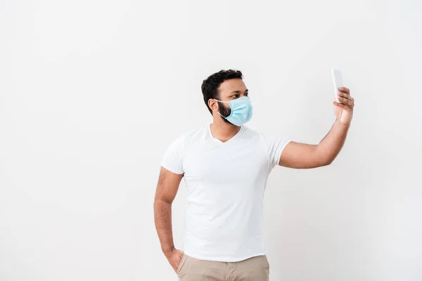 Афроамериканський чоловік в медичній масці стоїть з рукою в кишені і приймає селфі біля білої стіни — стокове фото
