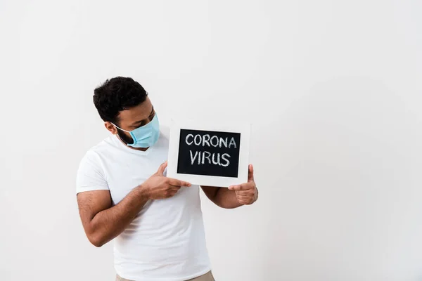 Homem americano africano em máscara médica azul olhando para quadro-negro com letras coronavírus perto da parede branca — Fotografia de Stock